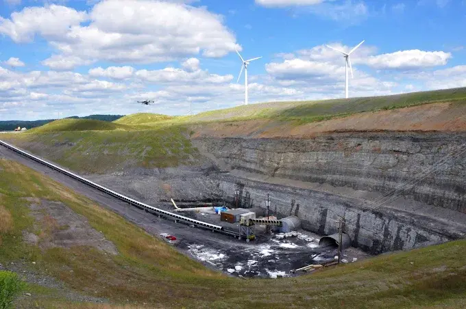 Drone flying over an open-pit mine site with a conveyor belt, excavation, and wind turbines in the background